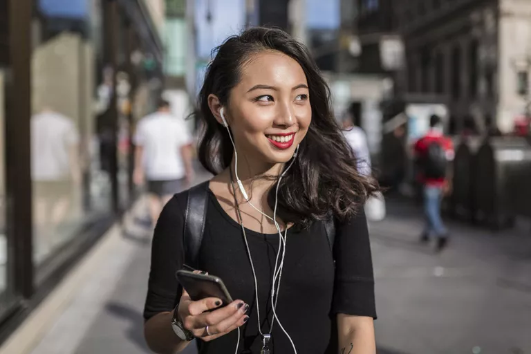 Smiling Asian woman listening to streaming music on her phone in a crowded city.