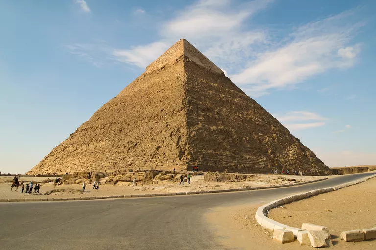 blue sky, large brown pyramid near road and small people and camel figures