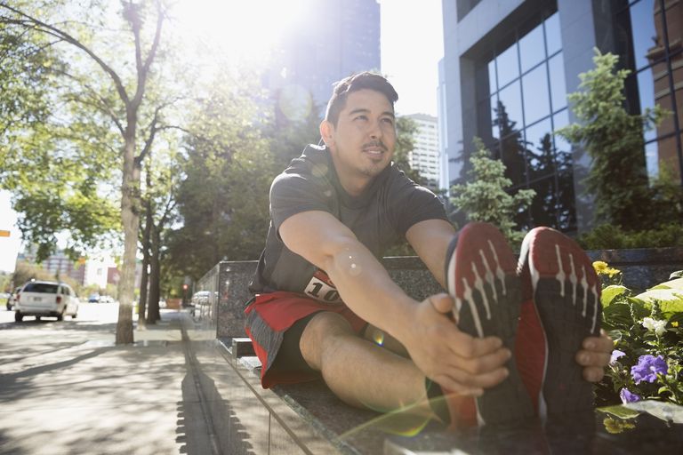 Male marathon runner stretching legs on sunny urban bench